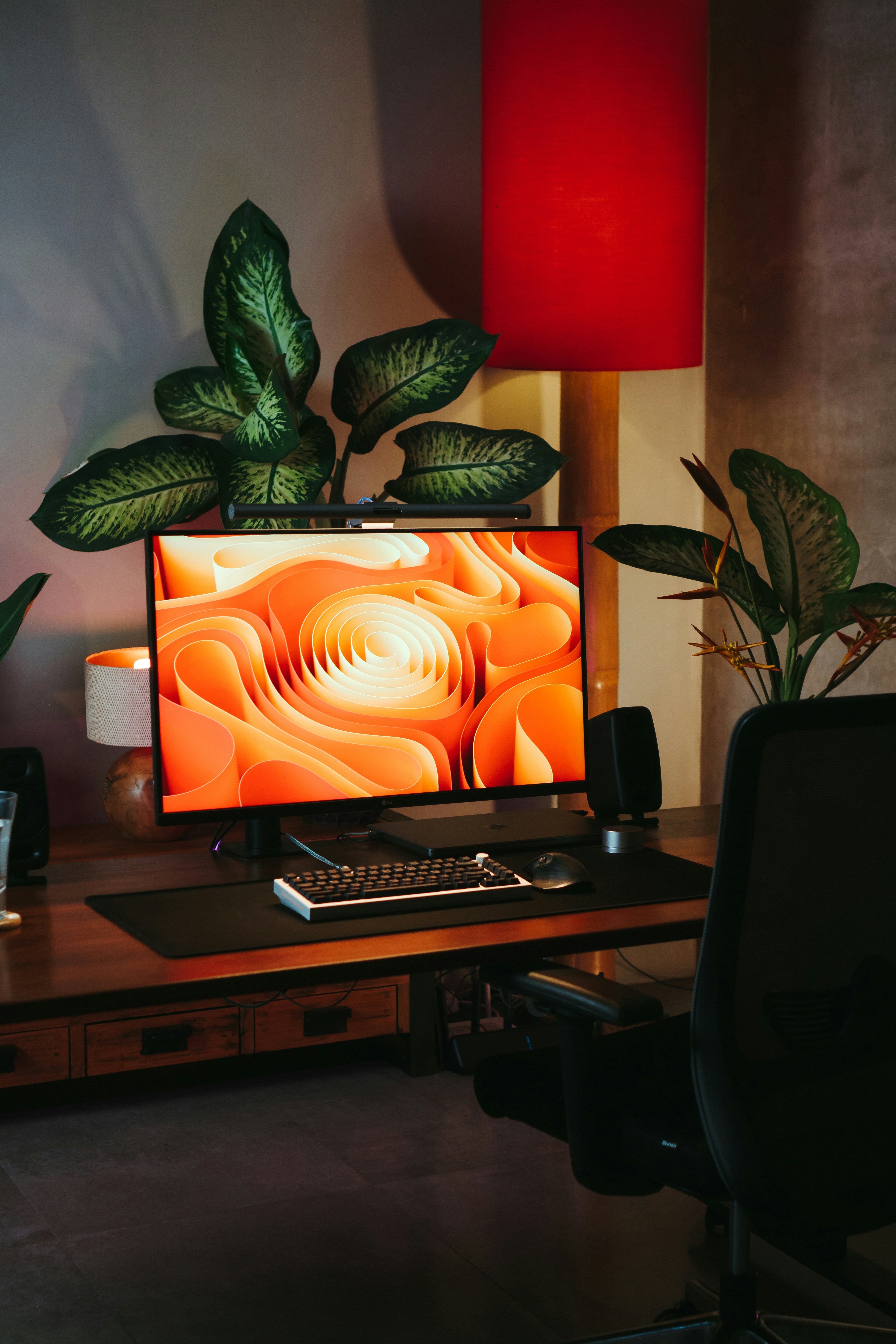 Modern workspace featuring a sleek desktop monitor displaying abstract orange art, surrounded by lush green plants and warm ambient lighting.