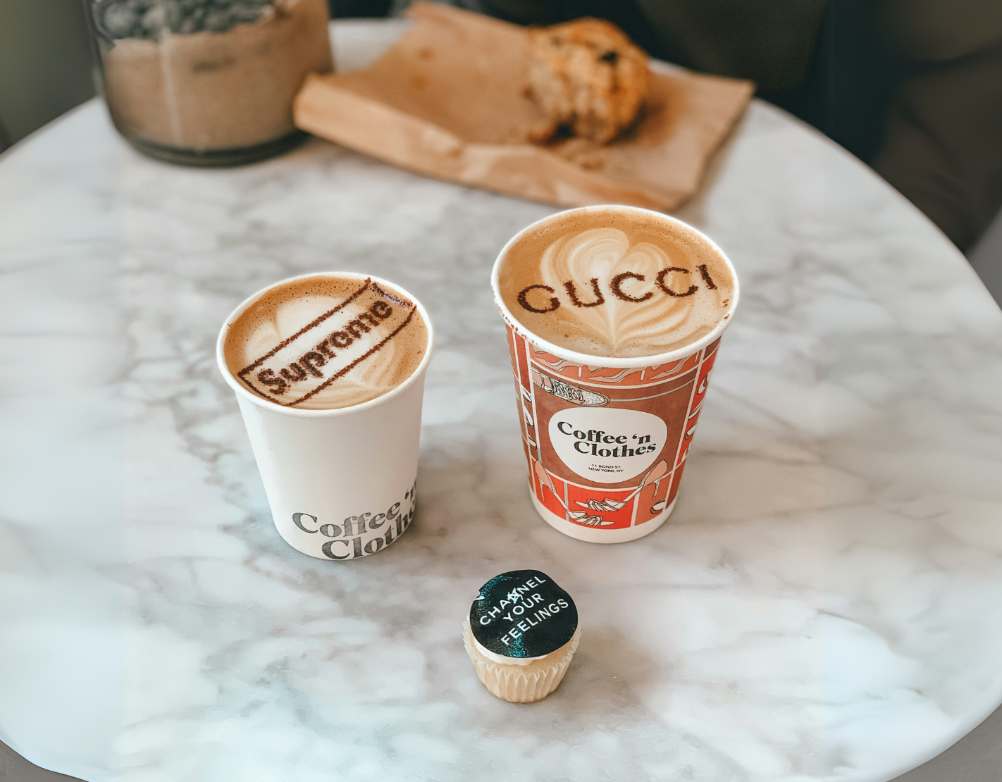 Two artisan coffee cups with branded latte art reading 'Supreme' and 'Gucci,' accompanied by a cupcake with the text 'Channel Your Feelings,' symbolizing luxury branding, creativity, and attention to detail.